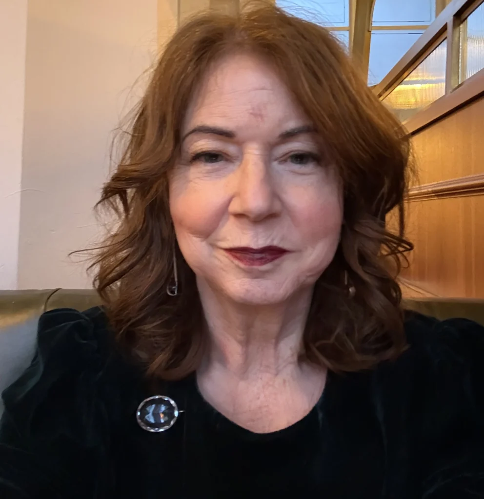 A close-up portrait shows a person with shoulder-length hair seated indoors, with a wooden panelled background and soft lighting.