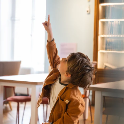 A child stands in a bright gallery space holding a magnifying glass and pointing upward, with tables, chairs, and a glass display case visible in the background.