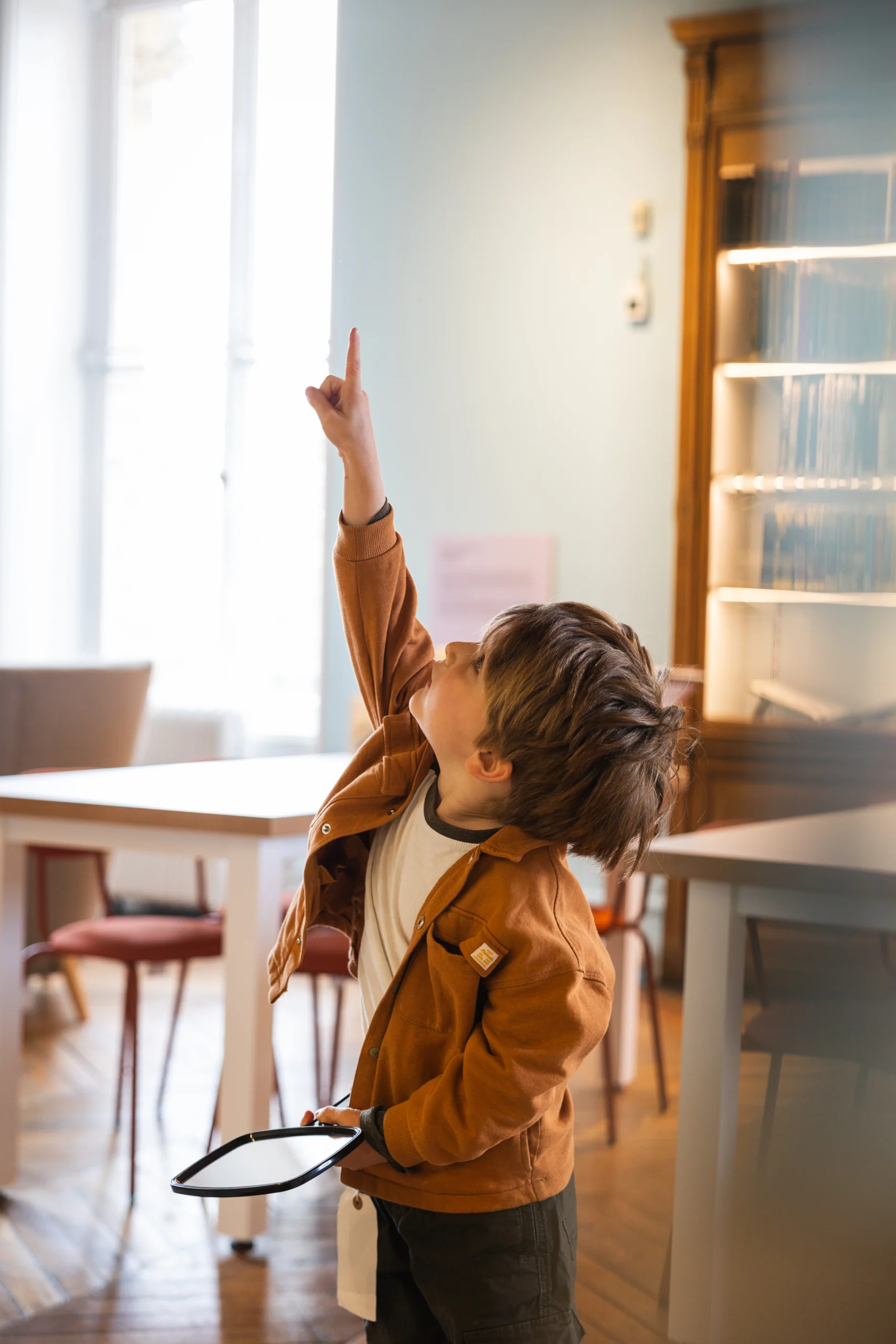 A child stands in a bright gallery space holding a magnifying glass and pointing upward, with tables, chairs, and a glass display case visible in the background.
