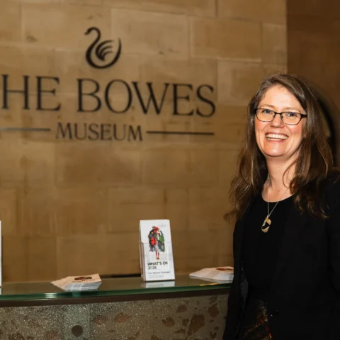 A visitor stands at a counter inside The Bowes Museum, with “The Bowes Museum” signage on a stone wall behind and leaflets displayed on the desk.