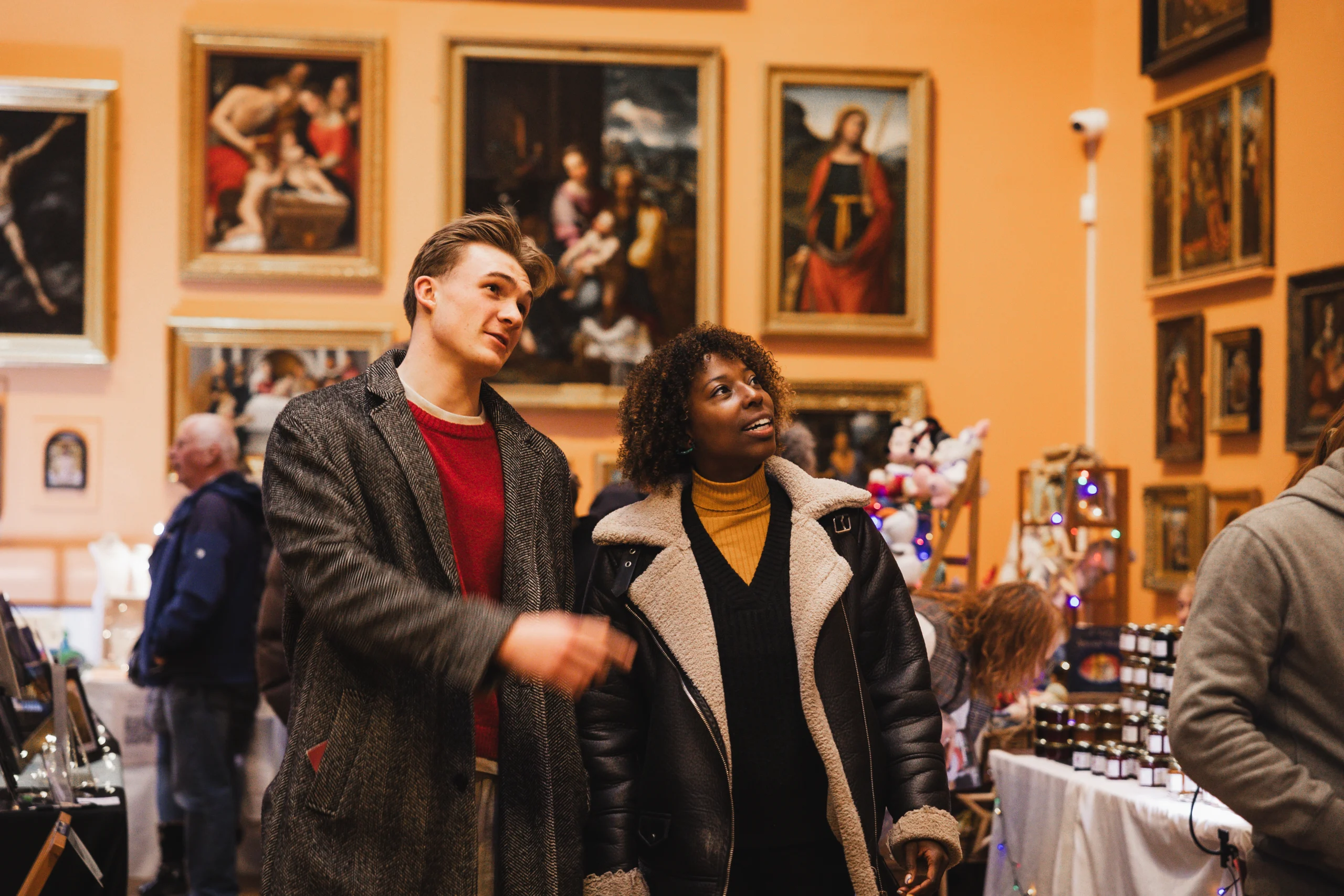 Two visitors stand in a gallery filled with framed paintings, looking upward while browsing a market-style display of goods on tables around them, with other visitors and stalls visible in the background.