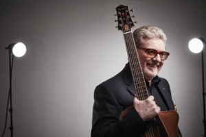 A musician holds a guitar over one shoulder while standing in front of a plain backdrop, with two bright studio lights positioned behind on either side.