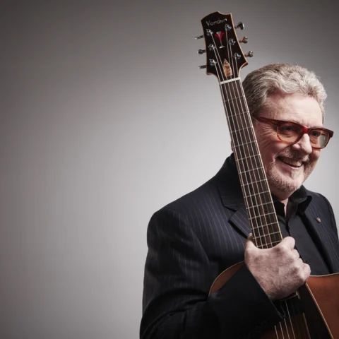 A musician holds a guitar over one shoulder while standing in front of a plain backdrop, with two bright studio lights positioned behind on either side.