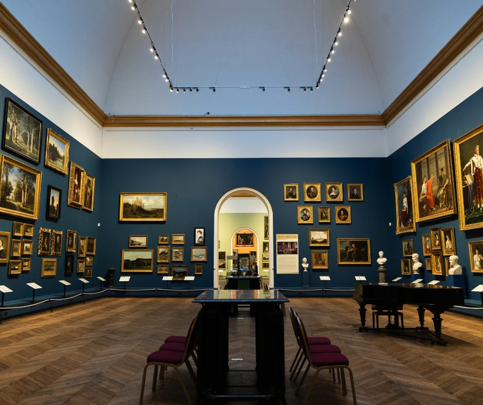 A large museum gallery with dark blue walls displays numerous framed paintings and several marble busts. Tables and chairs are arranged in the centre of the room, with an arched doorway leading to another gallery beyond.