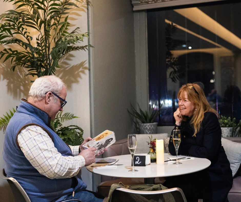 Two visitors sit at a small round table in a café, with glasses and tableware in front of them. One person looks at a booklet while the other sits opposite, surrounded by plants and soft lighting.