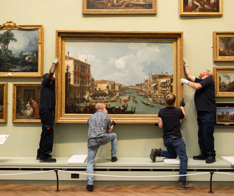 Four museum staff carefully position a large framed painting of a Venetian canal scene on a gallery wall, surrounded by other framed artworks. Several staff wear gloves while supporting the frame during installation.
