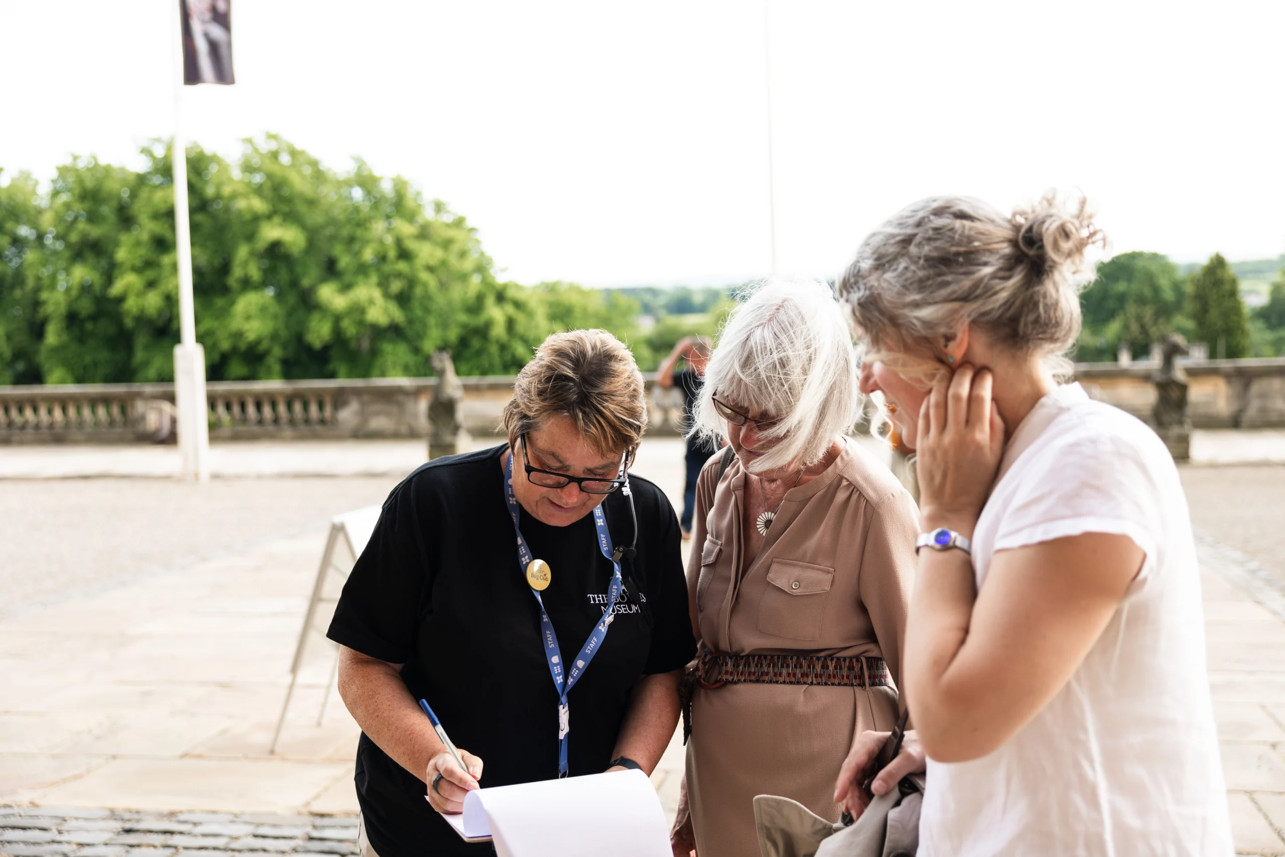 A museum staff member wearing a lanyard writes on a clipboard while speaking with two visitors outside a museum entrance. The three people stand together on a stone terrace, with balustrades, trees, and countryside views visible in the background.