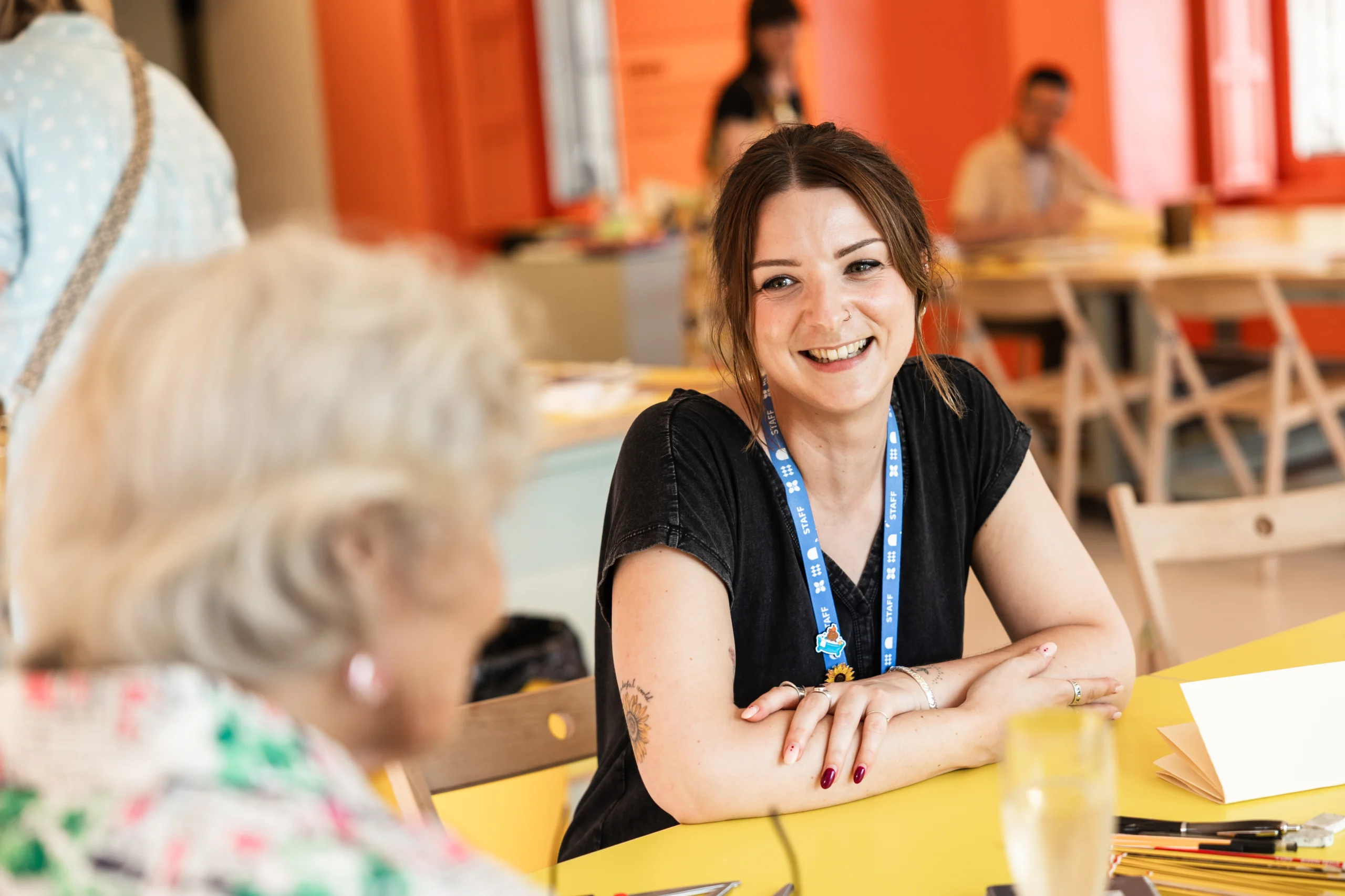 A museum staff member wearing a blue staff lanyard sits at a bright yellow table in a creative workshop space, talking with a visitor seated opposite. Pencils, paper, and drawing materials are spread across the table, while other participants work at tables in the background. The room features orange-red walls, large windows, and a light, open studio setting.