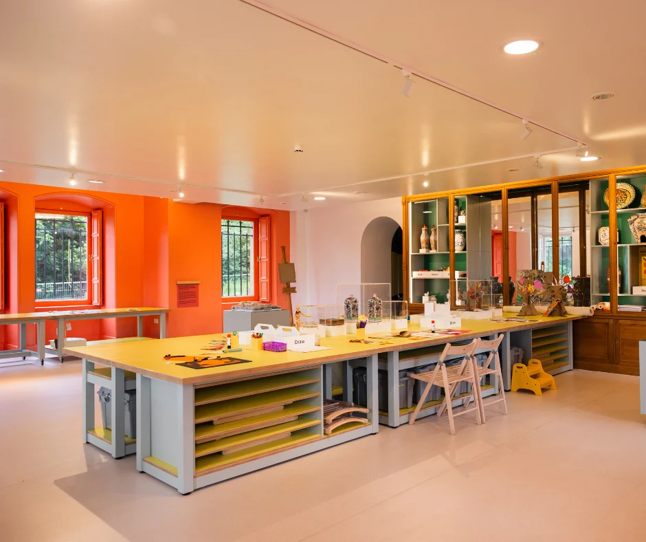 A bright creative learning space features large worktables covered with art materials and activity supplies. Orange walls, tall windows, storage units, and display cabinets filled with ceramics and objects surround the room.