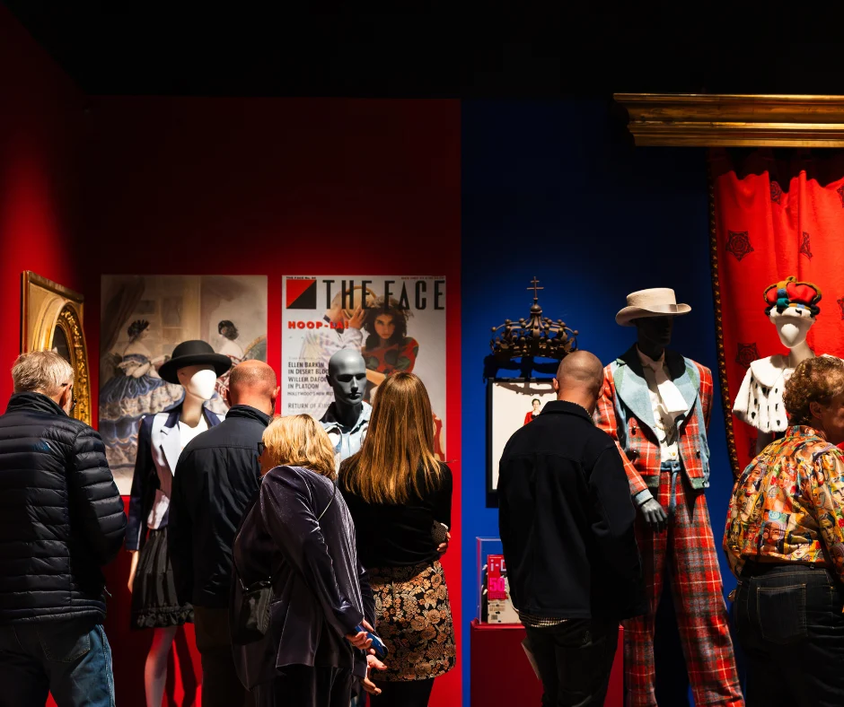Visitors view a fashion exhibition featuring mannequins dressed in Vivienne Westwood designs, displayed against bold red and blue walls alongside framed artworks, magazine covers, and interpretive panels.