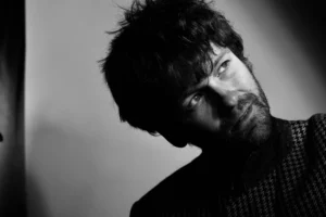 A black-and-white portrait shows a person with dark hair and a beard, photographed in dramatic side lighting. The person is angled slightly sideways and looking upward, wearing a textured patterned garment. Strong contrasts between light and shadow create a striking composition against a plain background.