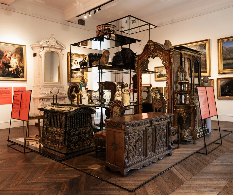 A gallery display showcases ornate antique furniture, ceramics, sculptures, and decorative objects arranged within a central open shelving structure, surrounded by framed paintings and interpretation panels.