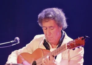 A musician sits on a stage playing an acoustic guitar while seated on a black stool. The performer wears a light-coloured suit, and purple stage lighting illuminates the scene. Microphones are positioned near the guitar, and the polished wooden stage floor reflects the coloured lights.