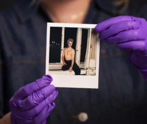 A person wearing purple gloves holds a Polaroid photograph showing a bare-chested figure posed by a window. The person holding the photo is softly out of focus in the background, with attention drawn to the small printed image in the foreground.
