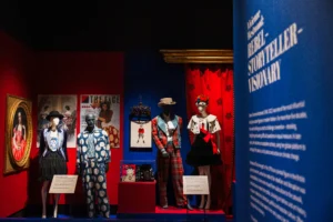 A fashion exhibition display features four mannequins dressed in Vivienne Westwood designs, including tailored jackets, patterned fabrics, tartan, and a crown-inspired outfit. The display is set against contrasting red and blue walls, surrounded by framed artworks, magazine covers, and exhibition panels. A large blue panel in the foreground includes the words “Rebel,” “Storyteller,” and “Visionary.”