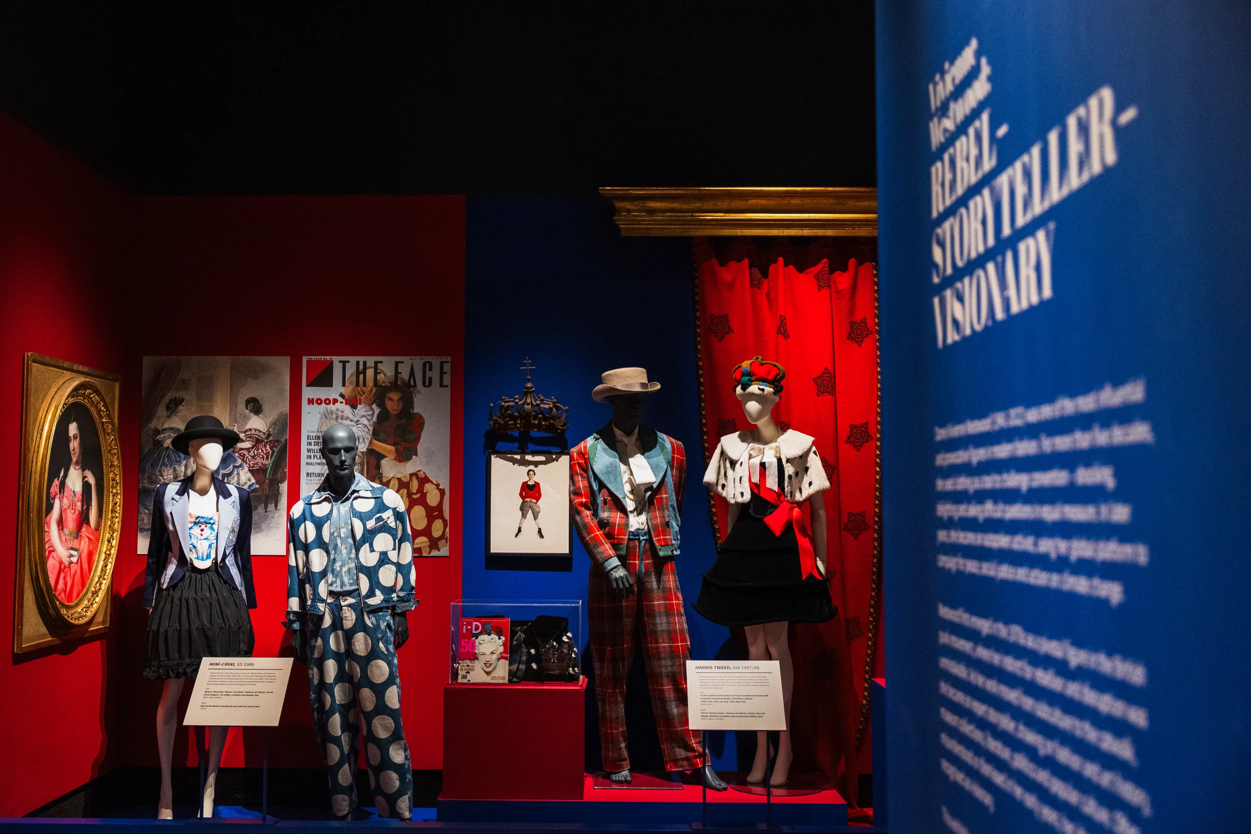 A fashion exhibition display features four mannequins dressed in Vivienne Westwood designs, including tailored jackets, patterned fabrics, tartan, and a crown-inspired outfit. The display is set against contrasting red and blue walls, surrounded by framed artworks, magazine covers, and exhibition panels. A large blue panel in the foreground includes the words “Rebel,” “Storyteller,” and “Visionary.”