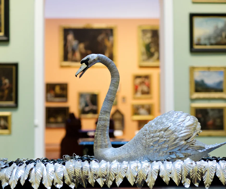 The Silver Swan automaton is displayed on an ornate base decorated with silver leaves. The swan's long neck and detailed feathered body are shown in profile, with framed paintings visible in the gallery beyond.