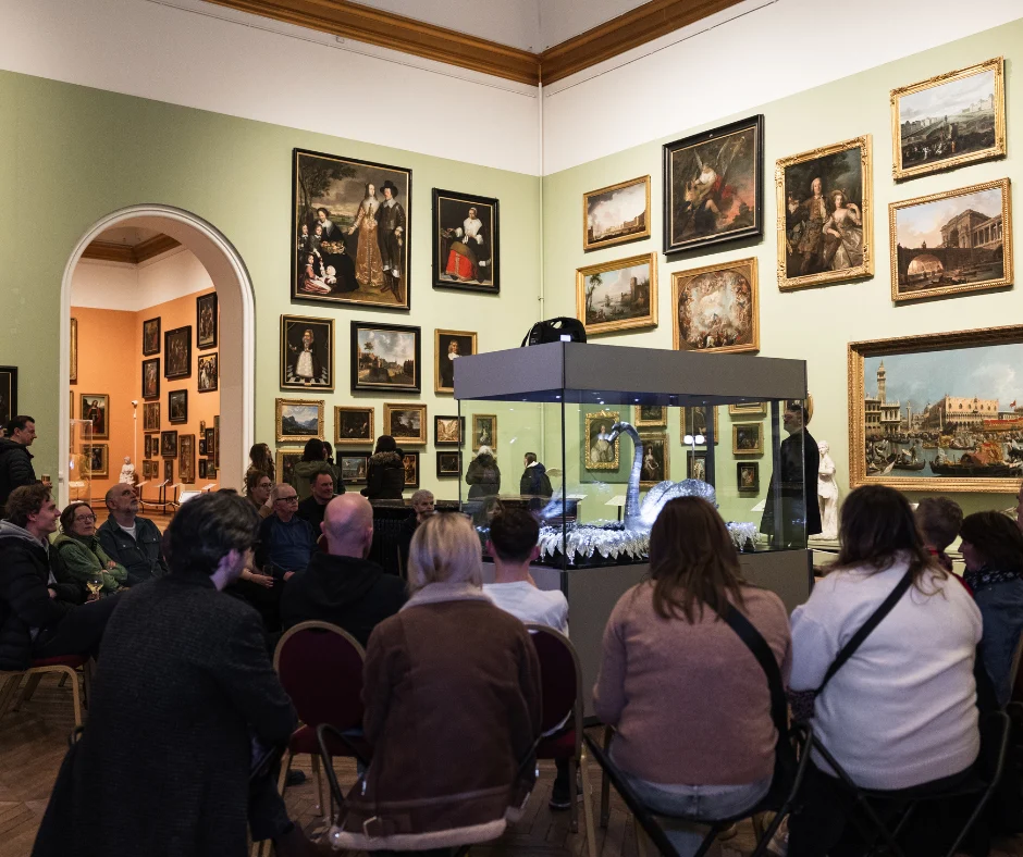 Visitors sit facing a large glass display case containing the Silver Swan installation during a gallery event. The room is filled with framed paintings on pale green walls, and rows of chairs are arranged around the display.
