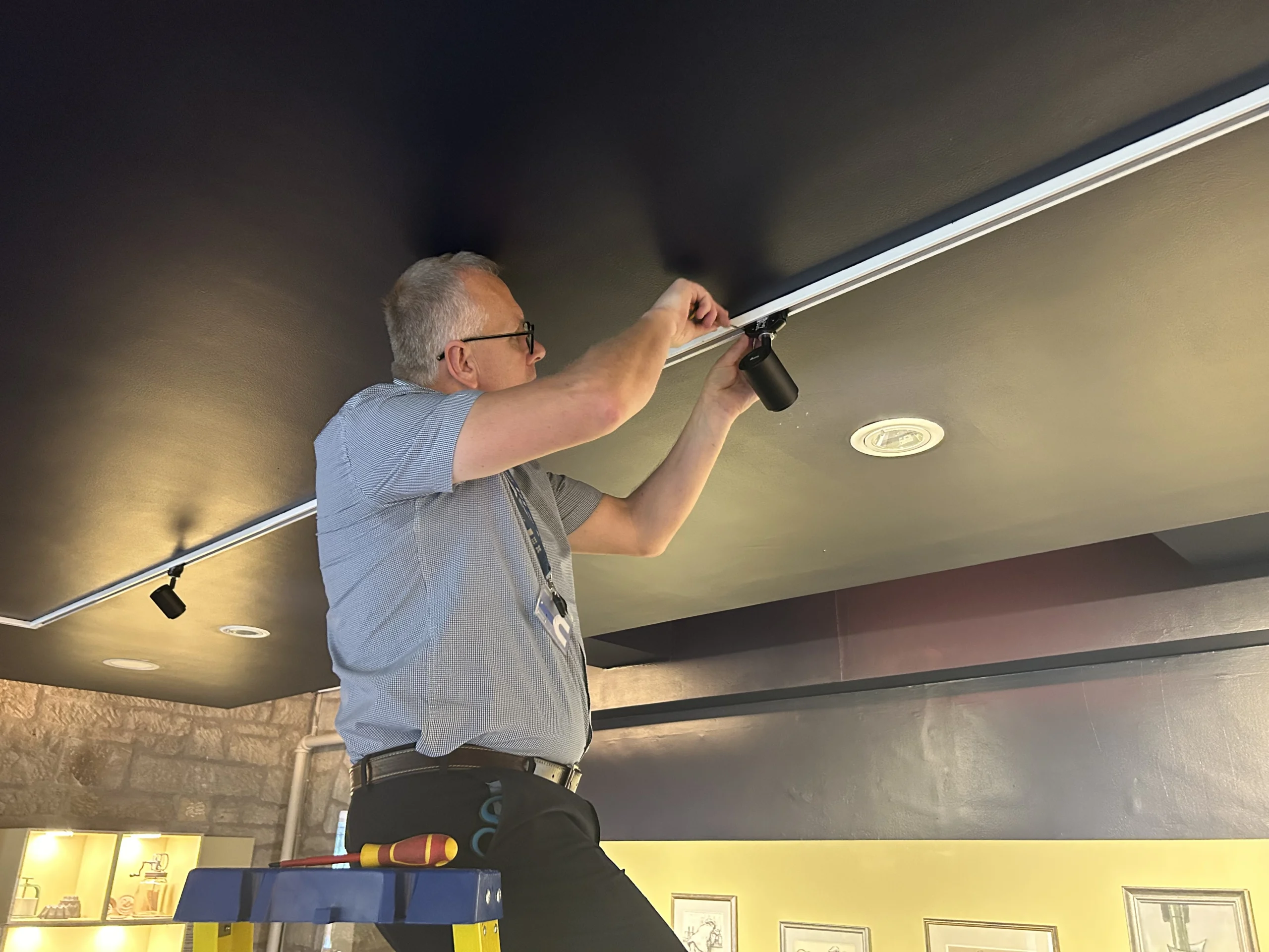 A museum staff member stands on a step ladder adjusting a black track-mounted spotlight attached to the ceiling. A toolbox and screwdriver rest on the platform below, while framed artworks and illuminated display cases are visible in the gallery space beneath a dark ceiling and stone walls.