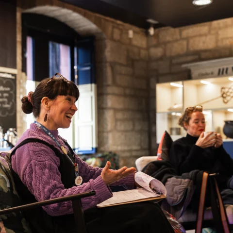 Two people sit in armchairs during a museum book club or discussion session in a café-style setting. One participant holds a notebook and speaks while seated, wearing a purple knitted jumper and a lanyard with badges. Another participant sits nearby holding a mug. Behind them, stone walls, display shelving with collection objects, and a chalkboard-style sign create a relaxed and welcoming museum environment.