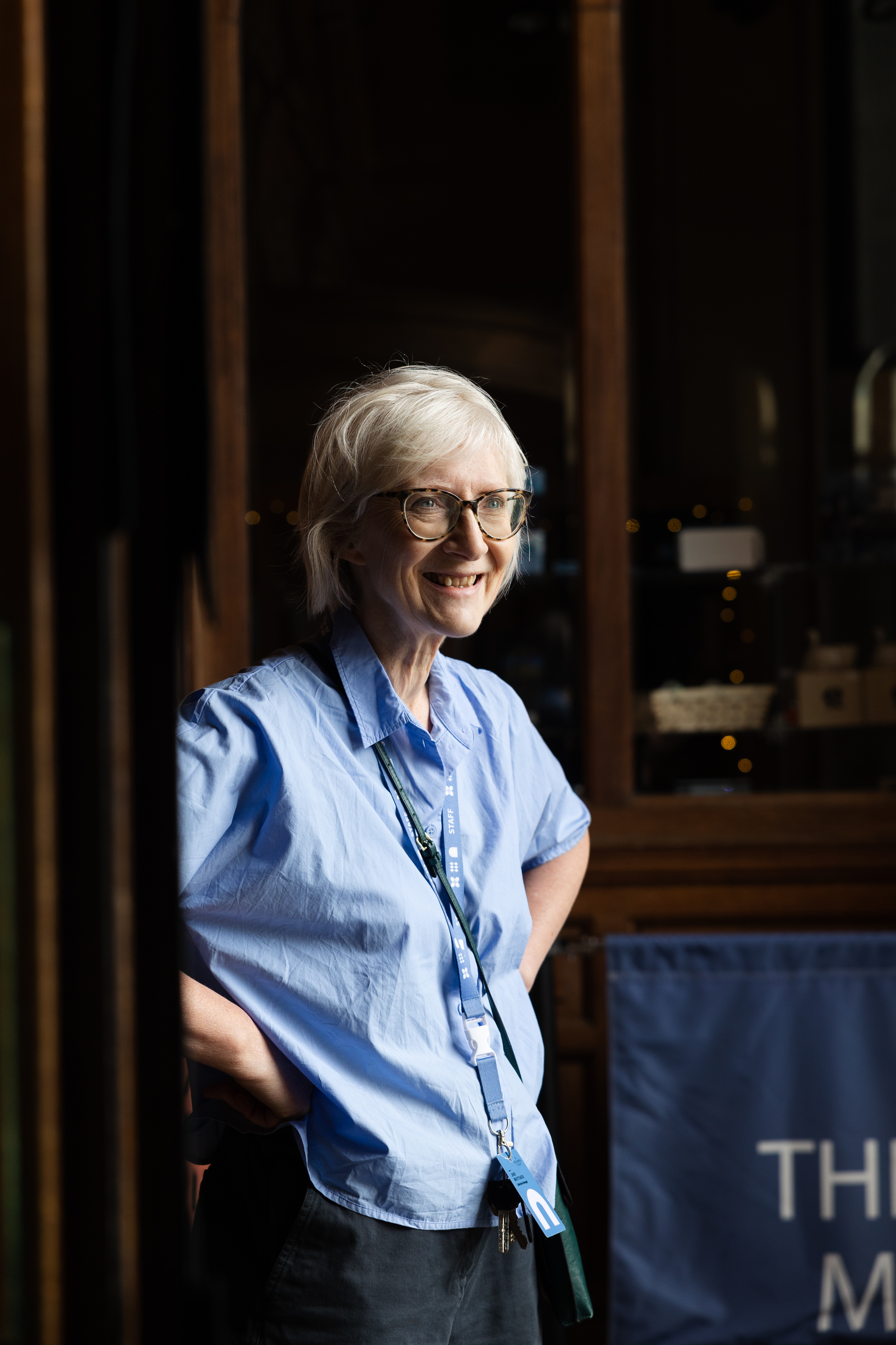 A museum staff member wearing glasses and a blue staff lanyard stands inside a historic interior with dark wooden display cabinets in the background. The person is positioned beside a doorway, wearing a light blue shirt and dark trousers. Warm lighting highlights the wood panelling and glass-fronted cabinets behind, while a Bowes Museum banner is partially visible in the foreground.