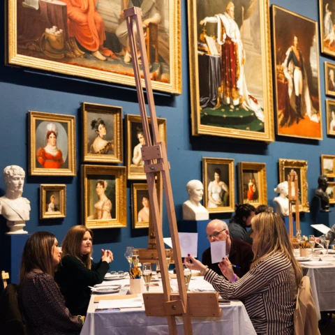 Visitors sit at tables set up with easels, pencils, paper, and drinks in a museum gallery during a creative evening event. The gallery’s dark blue walls are covered with framed paintings in ornate gold frames, while marble busts are displayed on plinths between the artworks. Participants are seated in small groups, talking and working on sketching activities beneath the surrounding paintings.