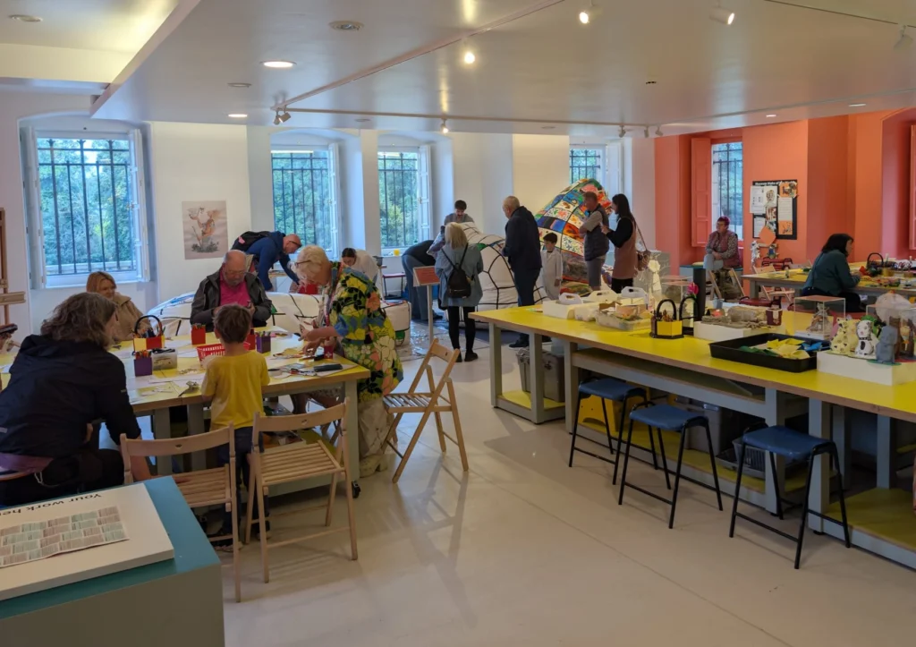 Photograph of a bright, spacious art workshop with people of various ages engaged in creative activities at tables. The room features large windows, white walls, yellow and orange accents, and displays of artwork and craft materials.