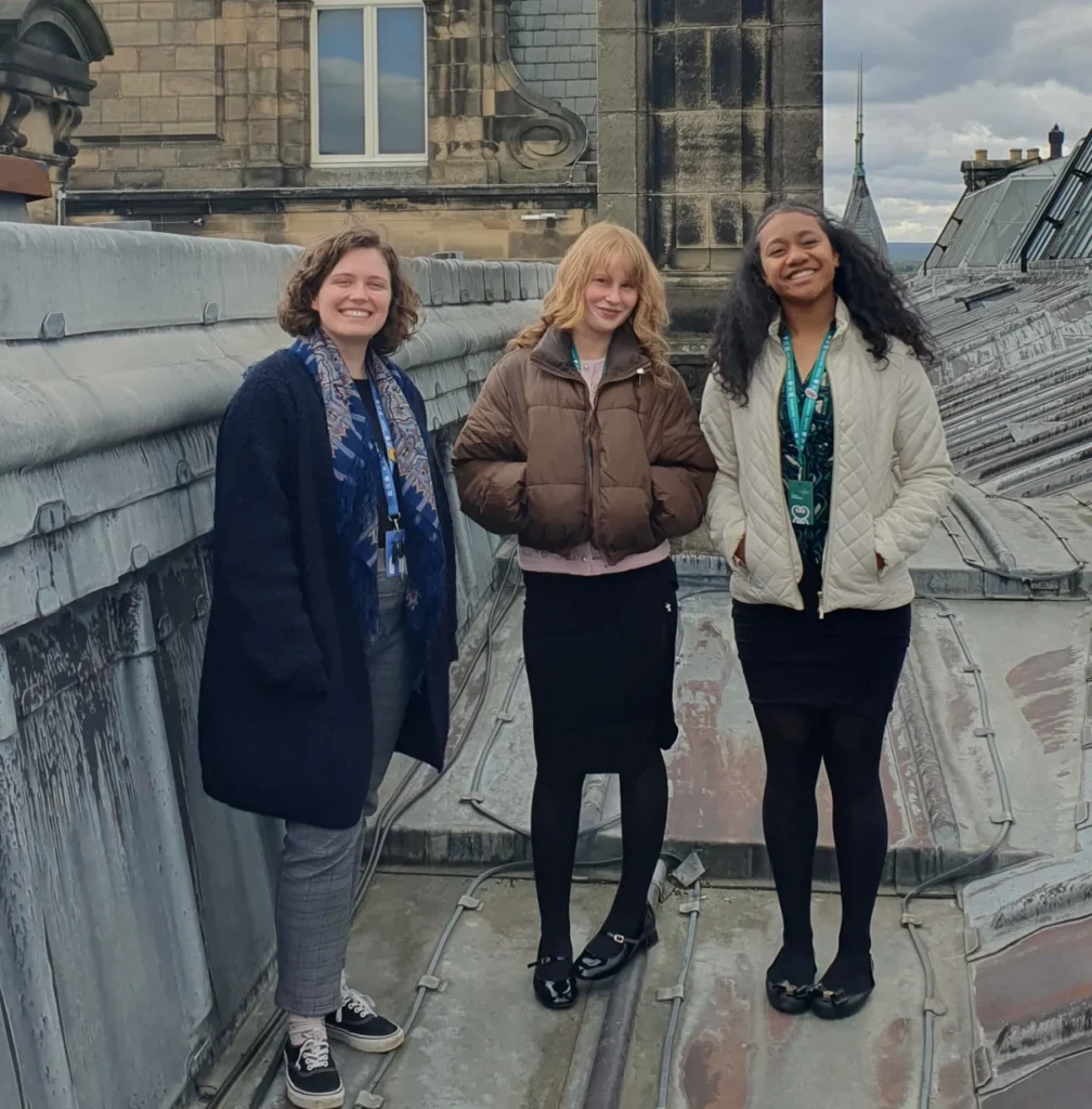 Three people stand on a lead-covered rooftop of a historic building, with ornate stone architecture, dormer windows, and chimneys visible around them. The group is positioned beside a large stone roof structure, with slate rooftops extending into the distance beneath a cloudy sky. Several people wear visitor or staff lanyards.