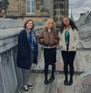 Three people stand on a lead-covered rooftop of a historic building, with ornate stone architecture, dormer windows, and chimneys visible around them. The group is positioned beside a large stone roof structure, with slate rooftops extending into the distance beneath a cloudy sky. Several people wear visitor or staff lanyards.