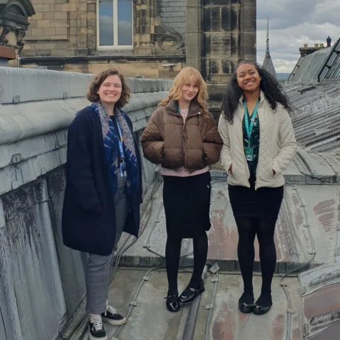 Three people stand on a lead-covered rooftop of a historic building, with ornate stone architecture, dormer windows, and chimneys visible around them. The group is positioned beside a large stone roof structure, with slate rooftops extending into the distance beneath a cloudy sky. Several people wear visitor or staff lanyards.