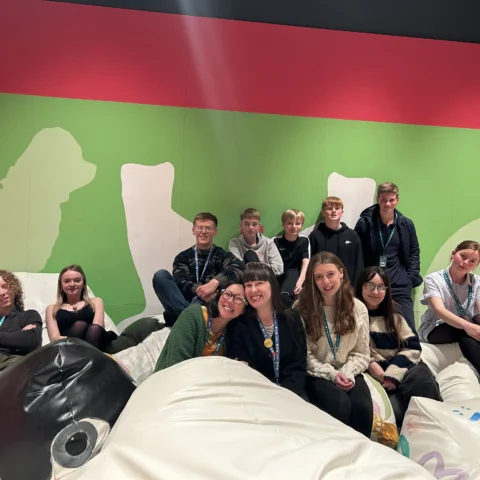 A group of young volunteers and museum staff sit together on large cushioned seating within a brightly coloured exhibition space. The backdrop features a vivid green wall with large pale silhouettes of animals and abstract shapes. The group wear visitor or volunteer lanyards and are gathered across oversized soft sculptural forms decorated with painted patterns, creating an informal seating area within the gallery.
