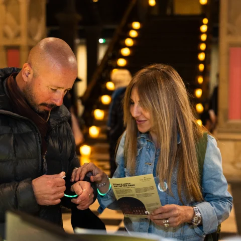 Two visitors stand in the grand entrance hall of The Bowes Museum while fastening glowing green wristbands onto their wrists. One visitor holds an event leaflet titled “Bowes Lates: Art After Dark”, while the other wears a dark jacket and scarf. The museum’s illuminated staircase rises behind them, lined with warm lights, and the historic interior features stone columns, arches, and a chequered floor. Other visitors and displays are visible in the softly lit background, suggesting an evening event atmosphere.