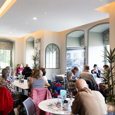 Visitors sit at tables in a bright museum café, eating, drinking, and talking together. Large arched windows with patterned blinds fill the space with natural light, while decorative wall panels, mirrors, and potted plants add detail to the interior. Tables are set with cups, glasses, and menus, and visitors are seated throughout the café enjoying refreshments in a relaxed setting.