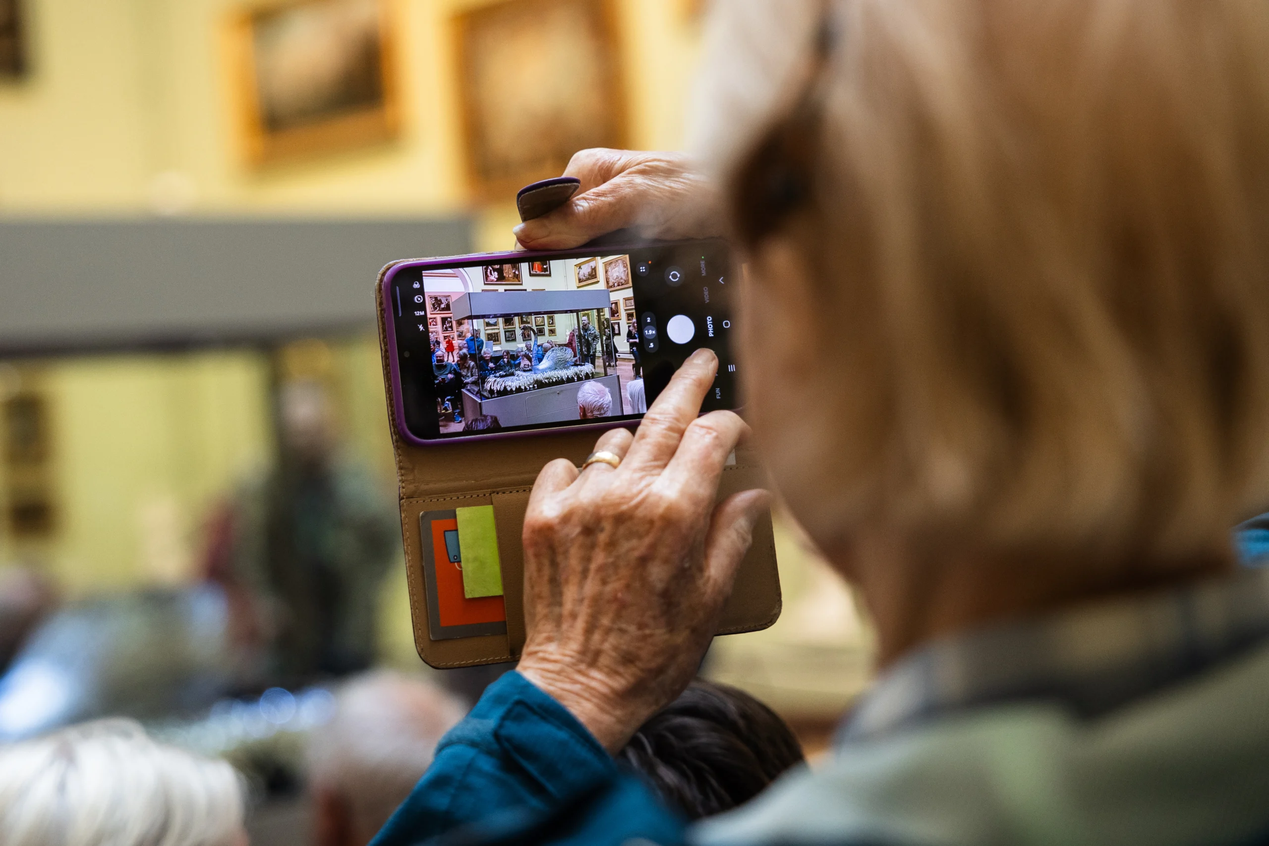 A visitor holds a smartphone to photograph the Silver Swan automaton inside a museum gallery. The phone screen shows the swan displayed in its glass case, surrounded by seated and standing visitors during a gallery event. Framed paintings hang on pale green walls in the background, while the visitor’s hands and phone case are visible in the foreground. The image captures the act of photographing the exhibition from within a crowd.