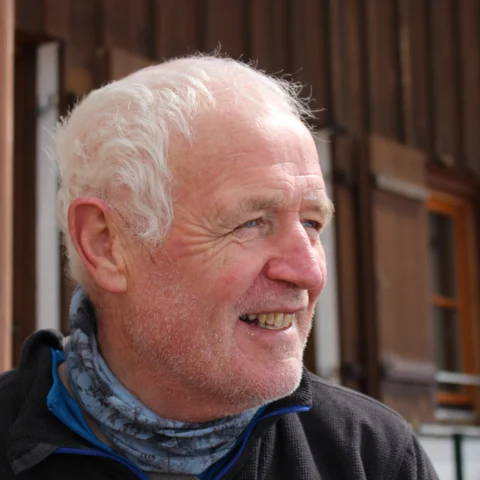 A portrait photograph shows an older person seated outdoors in front of a building with dark wooden cladding and large windows. The person has short white hair and wears a dark zip-up top with a patterned neck scarf. The image is cropped closely around the head and shoulders, with the background softly out of focus. Natural daylight highlights the facial features and texture of the wooden building behind.