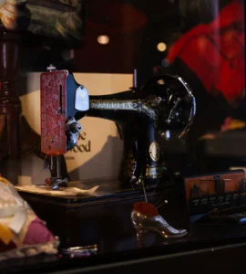 A museum display case features a vintage black Singer sewing machine decorated with gold detailing and mounted on a wooden base. Surrounding objects include a bright red Vivienne Westwood shopping bag with gold lettering, a small decorative shoe-shaped pincushion, sewing tools, fabric samples, and a cushion trimmed with lace and yellow ribbon. Reflections from the glass case and red exhibition displays appear in the background, linking the historic sewing equipment to the Vivienne Westwood fashion exhibition.