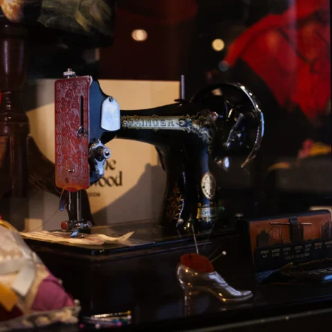 A museum display case features a vintage black Singer sewing machine decorated with gold detailing and mounted on a wooden base. Surrounding objects include a bright red Vivienne Westwood shopping bag with gold lettering, a small decorative shoe-shaped pincushion, sewing tools, fabric samples, and a cushion trimmed with lace and yellow ribbon. Reflections from the glass case and red exhibition displays appear in the background, linking the historic sewing equipment to the Vivienne Westwood fashion exhibition.