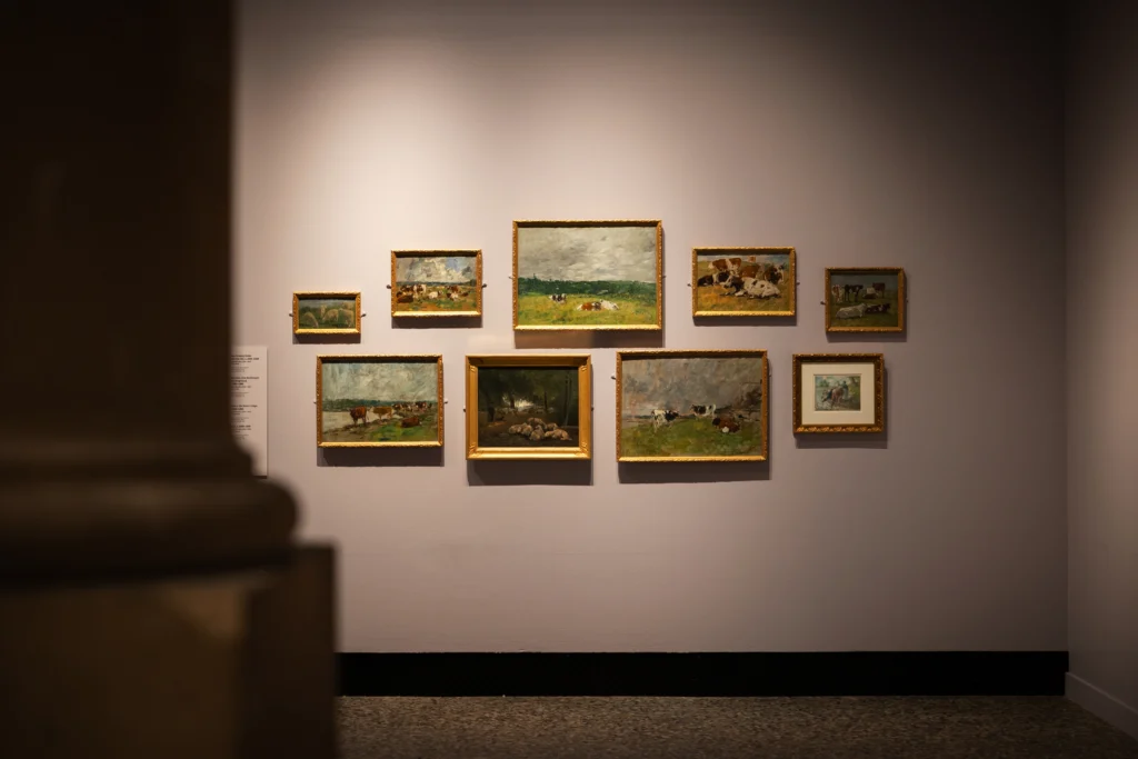 A gallery wall displays a group of small Impressionist paintings in gold frames. The artworks depict grazing cattle, sheep, rural landscapes, and pastoral scenes under expansive skies. Viewed through a doorway, the arrangement creates a focused display of countryside subjects illuminated by warm gallery lighting.