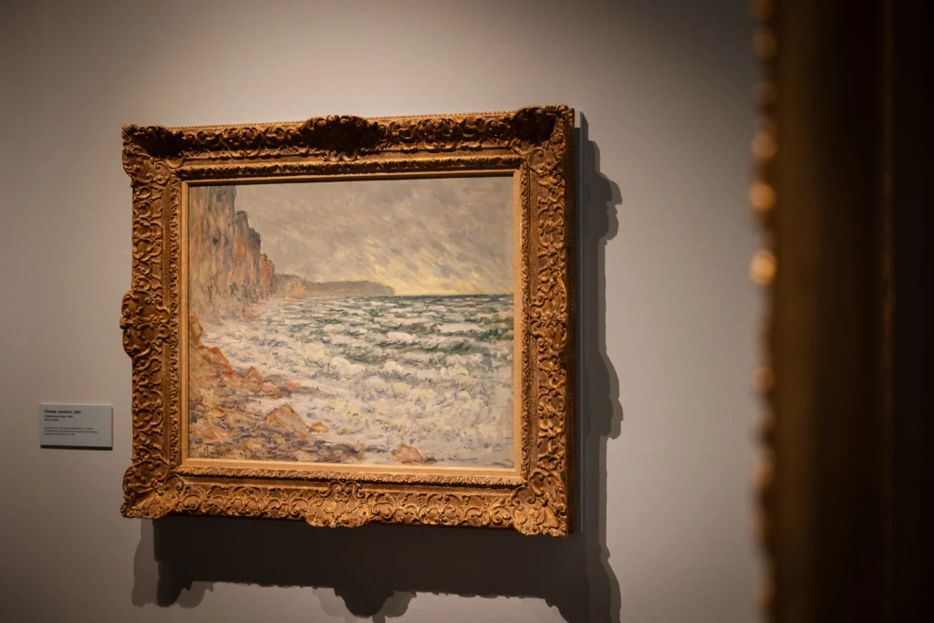 An Impressionist seascape painting hangs in an elaborate gilded frame on a gallery wall. The artwork depicts waves breaking against a rocky shoreline beneath a light-filled sky, with cliffs receding into the distance. Soft brushstrokes in pale blues, creams, and pinks capture the movement of the sea and changing light.