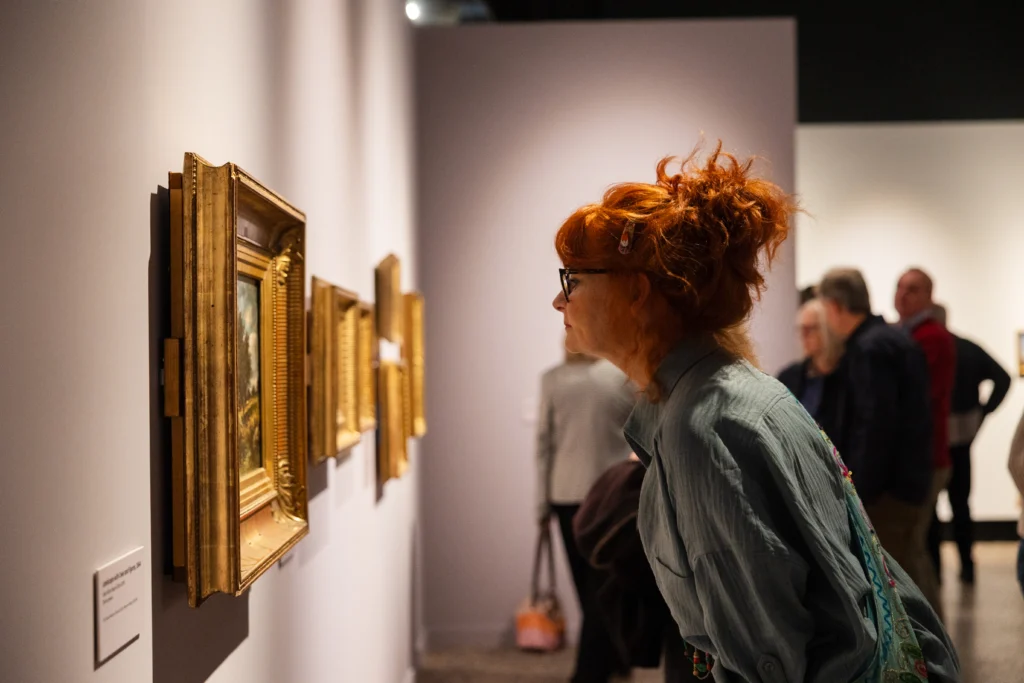 A visitor leans forward to closely examine a framed painting in a gallery. Several gold-framed artworks hang in a row along the pale wall, while other visitors stand further back in the exhibition space. Warm spotlighting illuminates the paintings and creates a focused viewing environment.