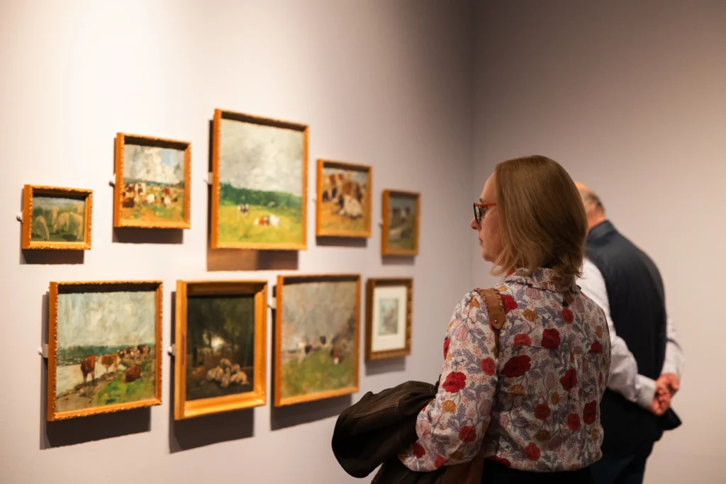 A visitor stands in a gallery looking at a wall display of small Impressionist paintings in ornate gold frames. The artworks depict pastoral scenes, grazing animals, figures in fields, and landscapes under expansive skies. Other visitors are visible in the background, while soft gallery lighting highlights the paintings against a pale wall.