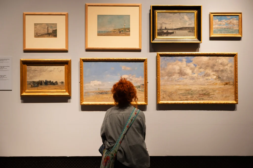 A visitor with bright red hair stands facing a gallery wall filled with framed seascapes and coastal scenes. The paintings, displayed in gold and wooden frames, depict beaches, boats, harbours, and expansive skies in soft Impressionist colours. The visitor is positioned centrally, studying the artworks from close range.