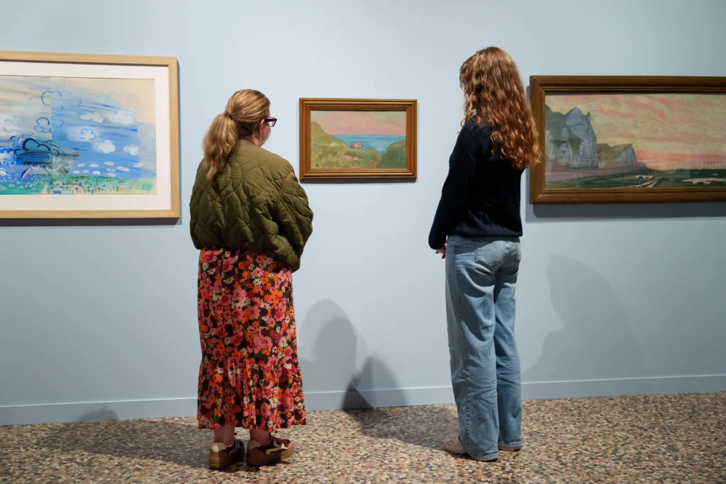Two visitors stand in a gallery viewing a display of artworks hung on pale blue walls. Between them is a small landscape painting showing a pink-roofed cottage overlooking the sea, while larger paintings on either side depict coastal scenes in soft pastel colours. One artwork features sweeping blue brushstrokes and cloud-like forms, while another shows cliffs and shoreline at sunset. The visitors stand quietly facing the paintings, with the gallery’s terrazzo floor and minimalist display creating a calm exhibition setting.