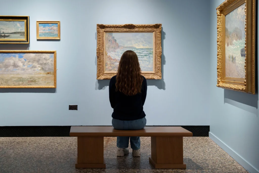 A visitor sits on a wooden bench in a gallery, facing a wall of Impressionist paintings displayed against pale blue walls. At the centre is a large seascape in an ornate gilded frame, flanked by smaller coastal and landscape paintings. The visitor is viewed from behind, seated quietly while studying the artworks in the softly lit exhibition space.